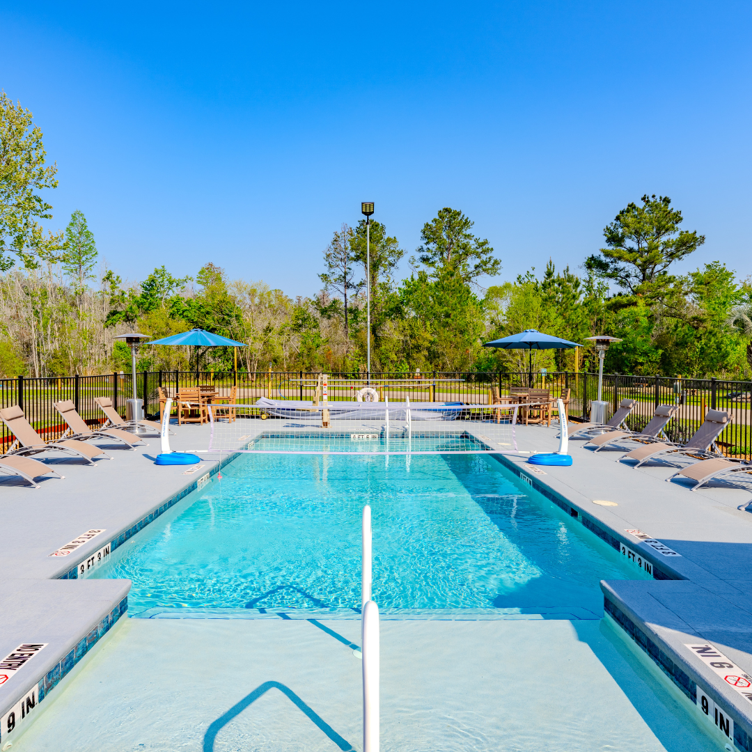 Wide shot of the outdoor pool at Lake City RV Resort, there is a net over the middle of the pool for volleyball. The pool is surrounded by lounging chairs and two umbrellas in the far corners. The pool is gated.